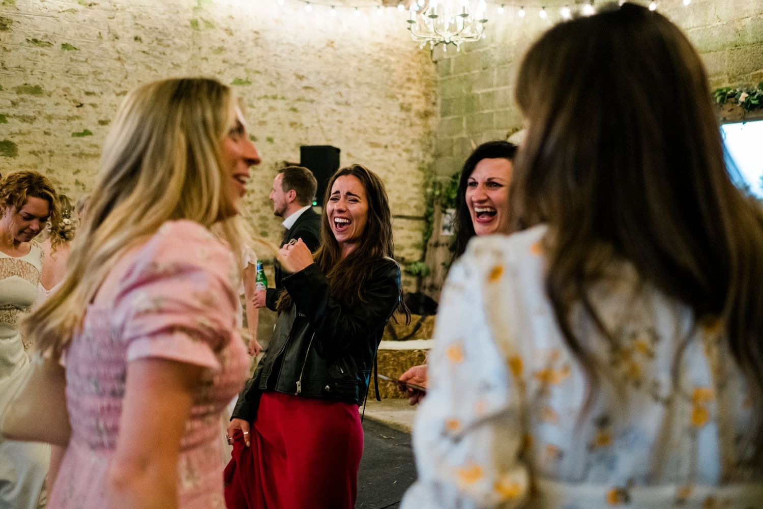 Wedding guests laughing and dancing together at a lively reception in a stone-walled venue.