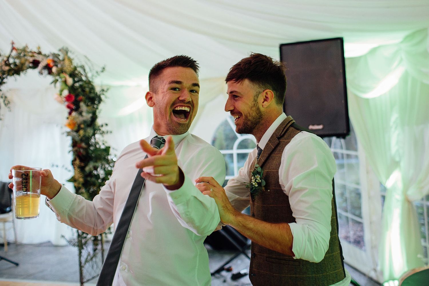 Two guests laugh and celebrate together at a wedding reception under floral decorations.