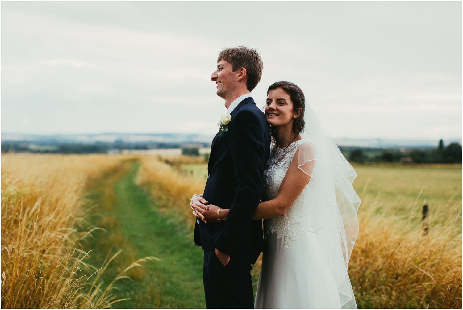 Newlyweds sharing intimate moment in rustic field with rolling hills in distance.