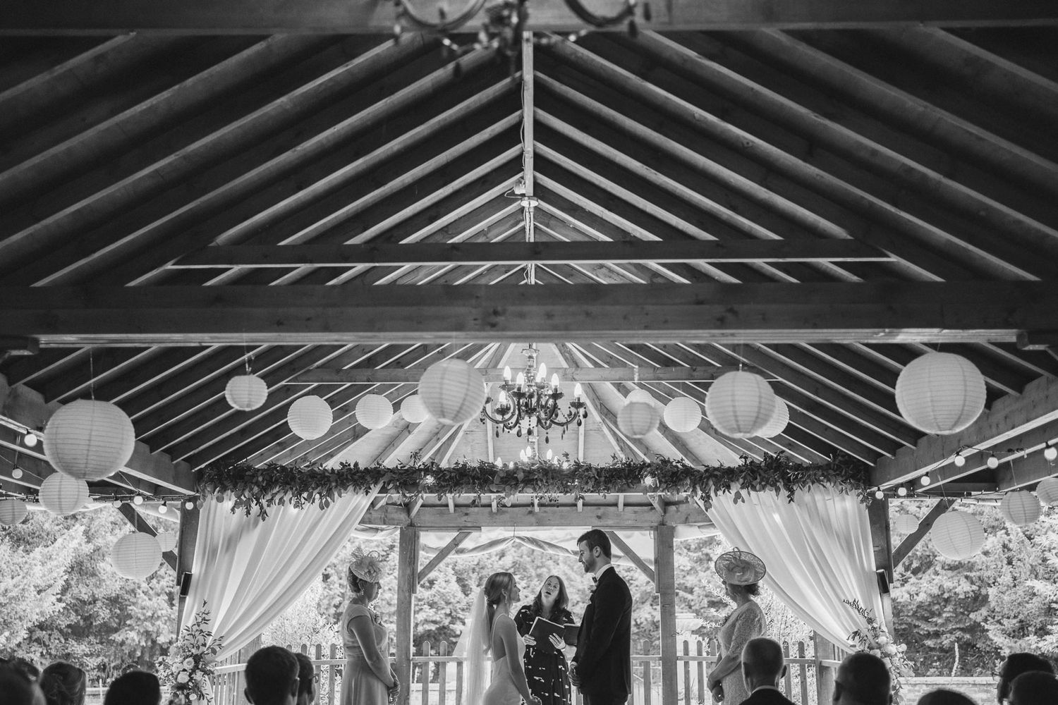 Black and white photo of a wedding ceremony in a barn with exposed wooden beams and hanging lanterns.