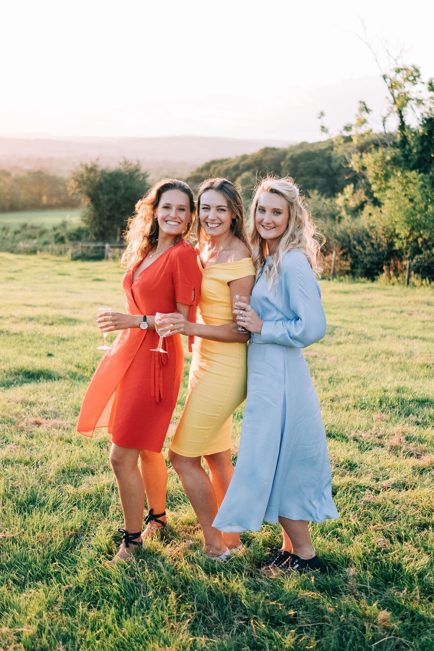 Bridesmaids in colorful retro dresses pose playfully in sunny field.