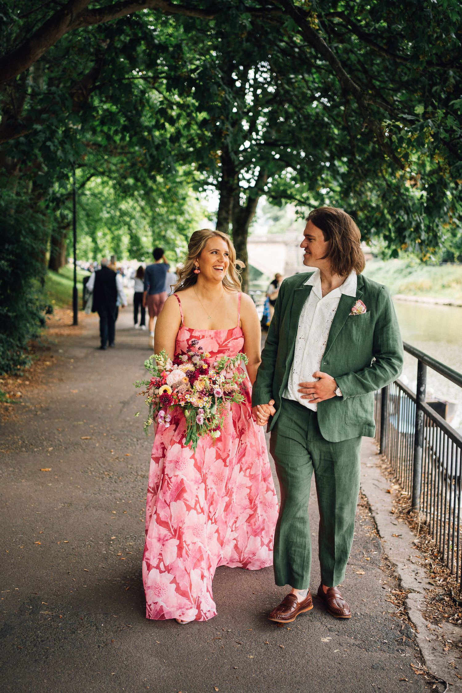 Wedding couple walking together outdoors with one wearing a vibrant pink floral dress and the other in a green suit.