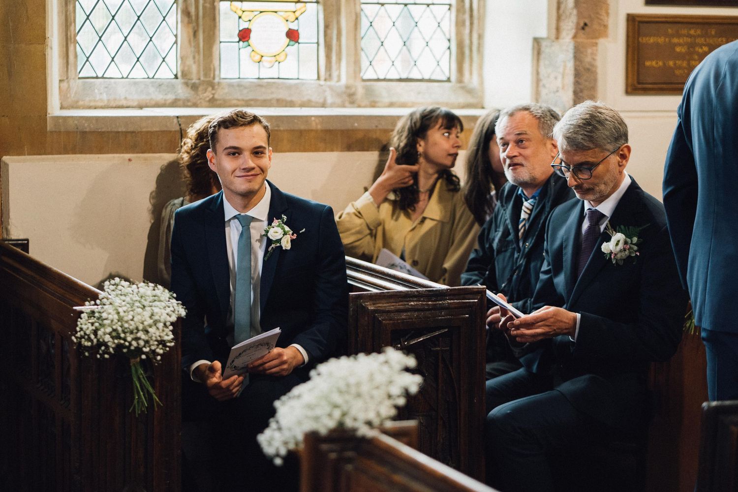 Wedding guests seated in wooden church pews with sunlight streaming through windows.