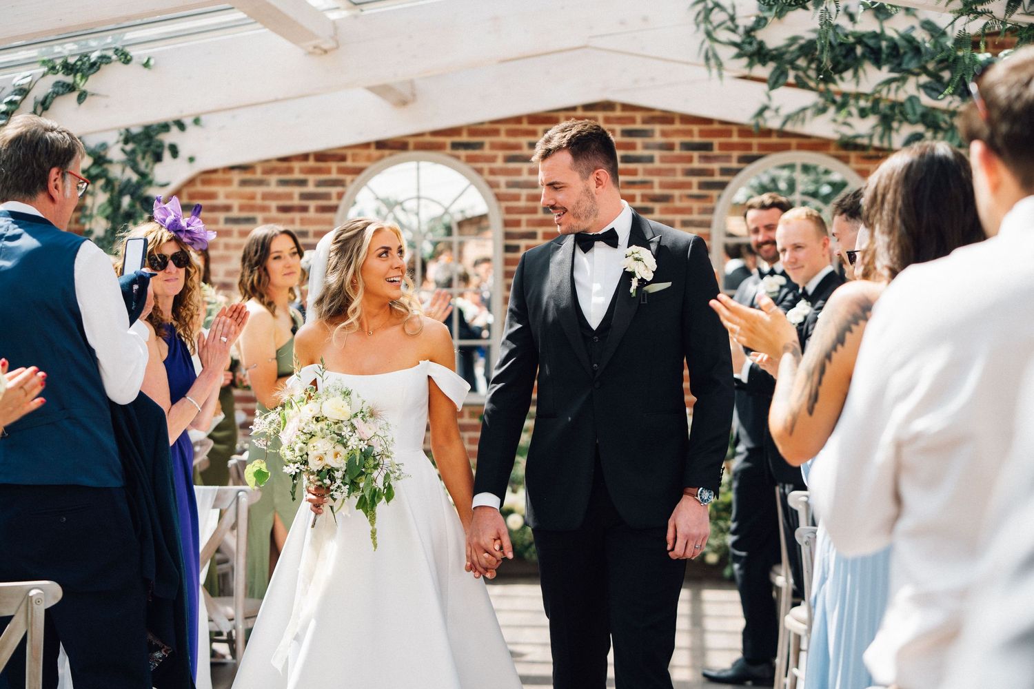 Newlyweds walk down the aisle at a brick-walled venue while guests celebrate with confetti and applause.