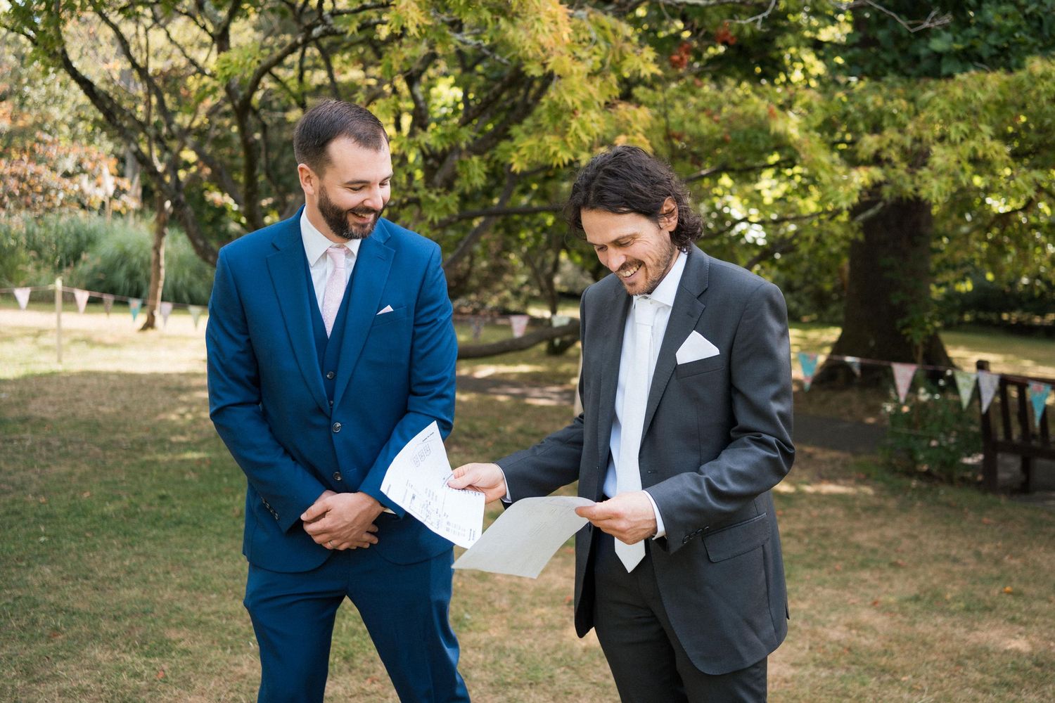 Two men in suits reviewing documents outdoors at a garden wedding ceremony.