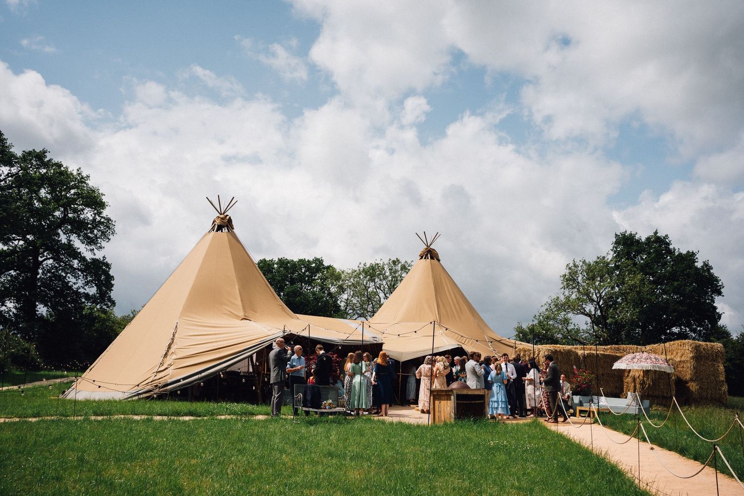 Two large tipi tents set up outdoors on green grass for a wedding event under a partly cloudy blue sky.