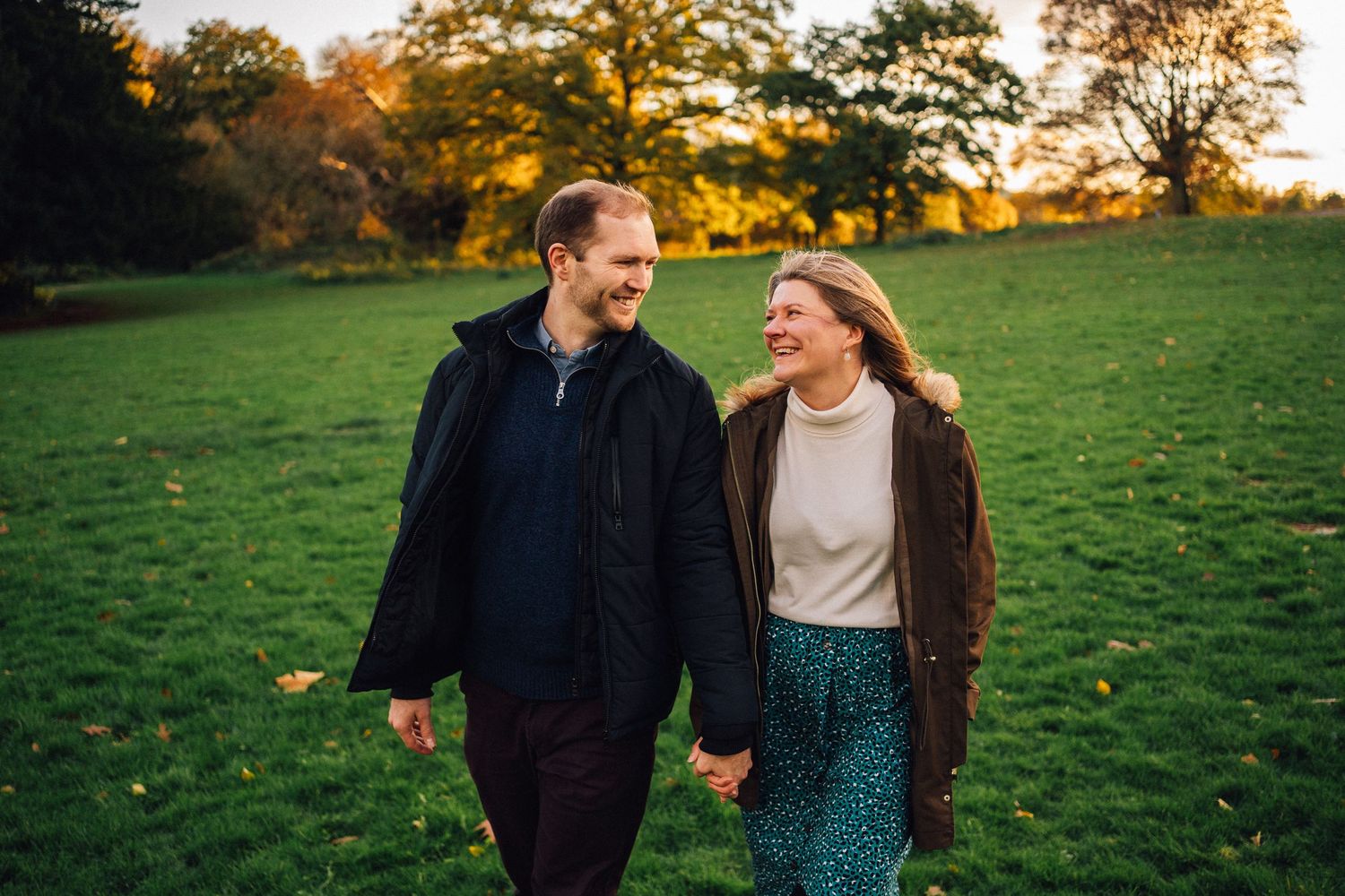 Two people strolling together on green grass during sunset with trees in the background.