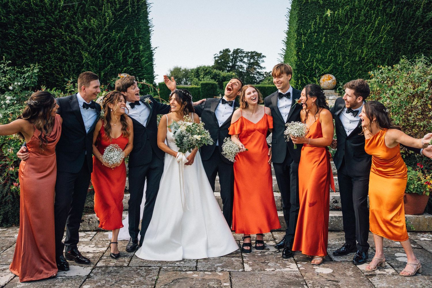 Bridesmaids in vibrant orange and rust-colored dresses pose together in an outdoor garden setting.