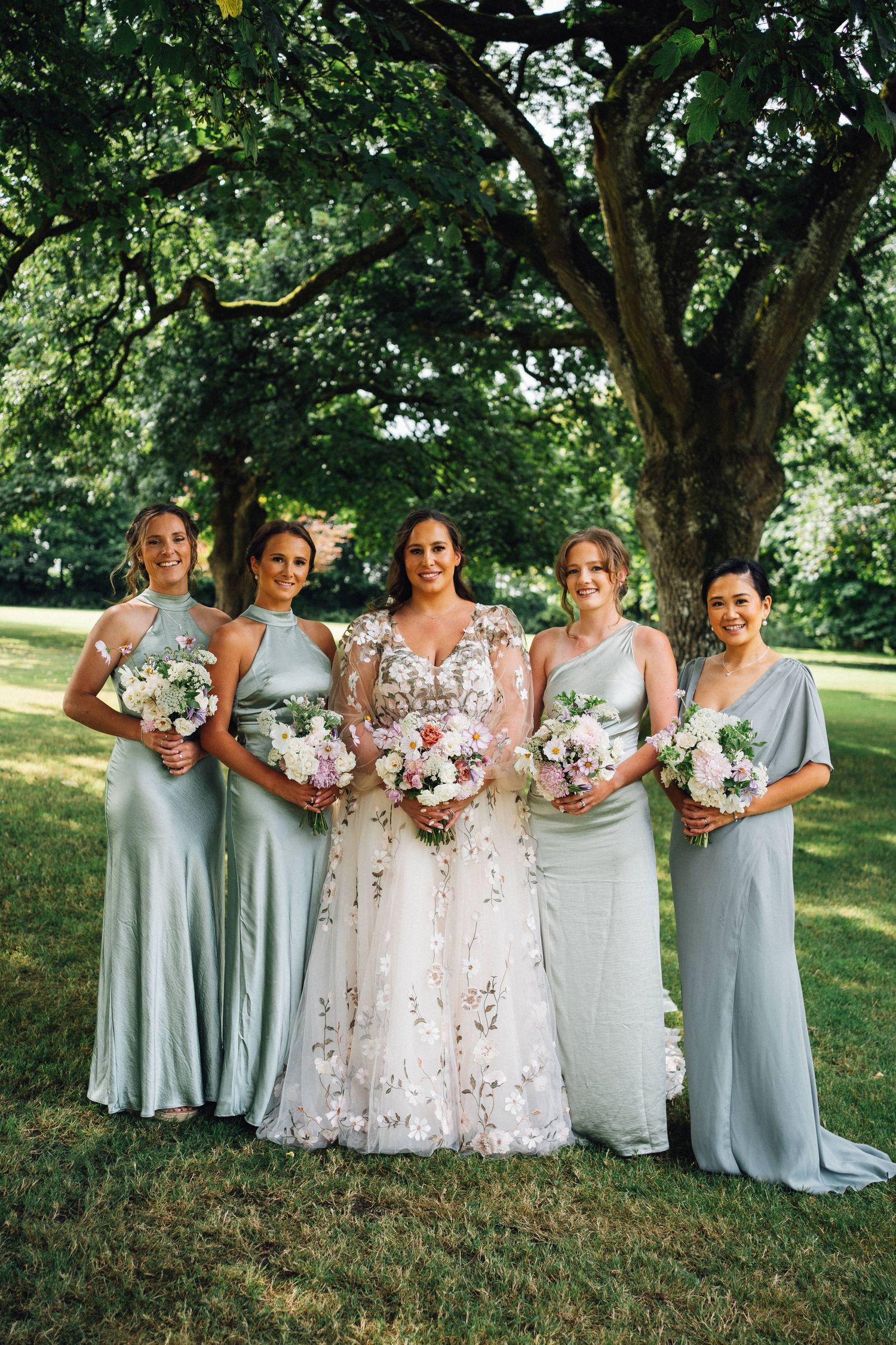 Wedding party in pale blue dresses poses under a large tree.