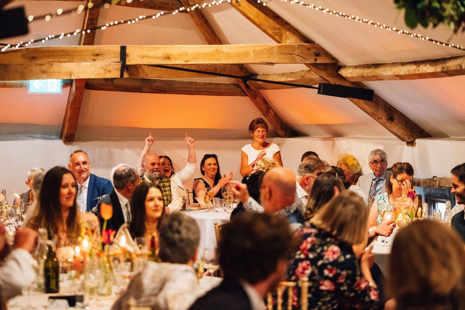 Wedding reception celebration with guests cheering under string lights in a rustic venue.