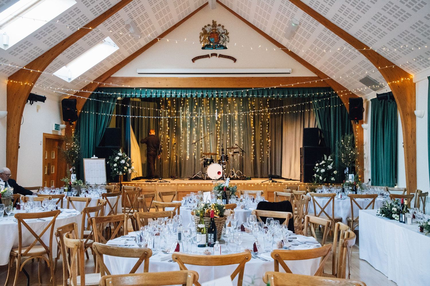 Elegant wedding reception hall with fairy lights, green drapes, wooden chairs, and floral centerpieces on white-clothed tables.