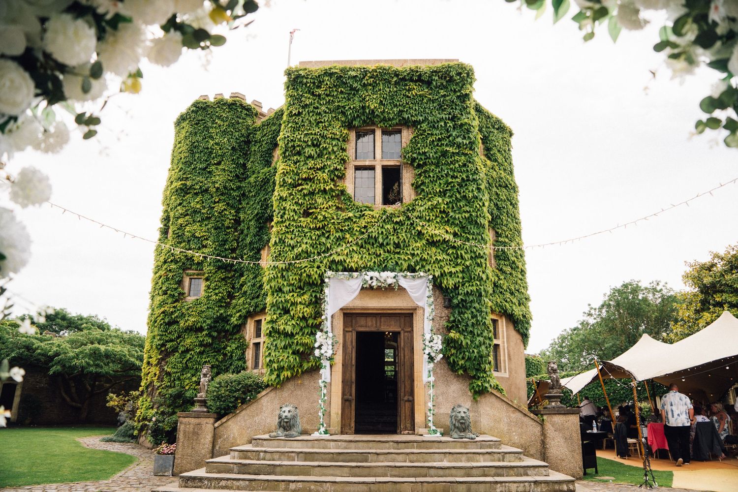 Ivy-covered stone wedding venue with white floral decorations, steps leading to entrance, surrounded by lush greenery.