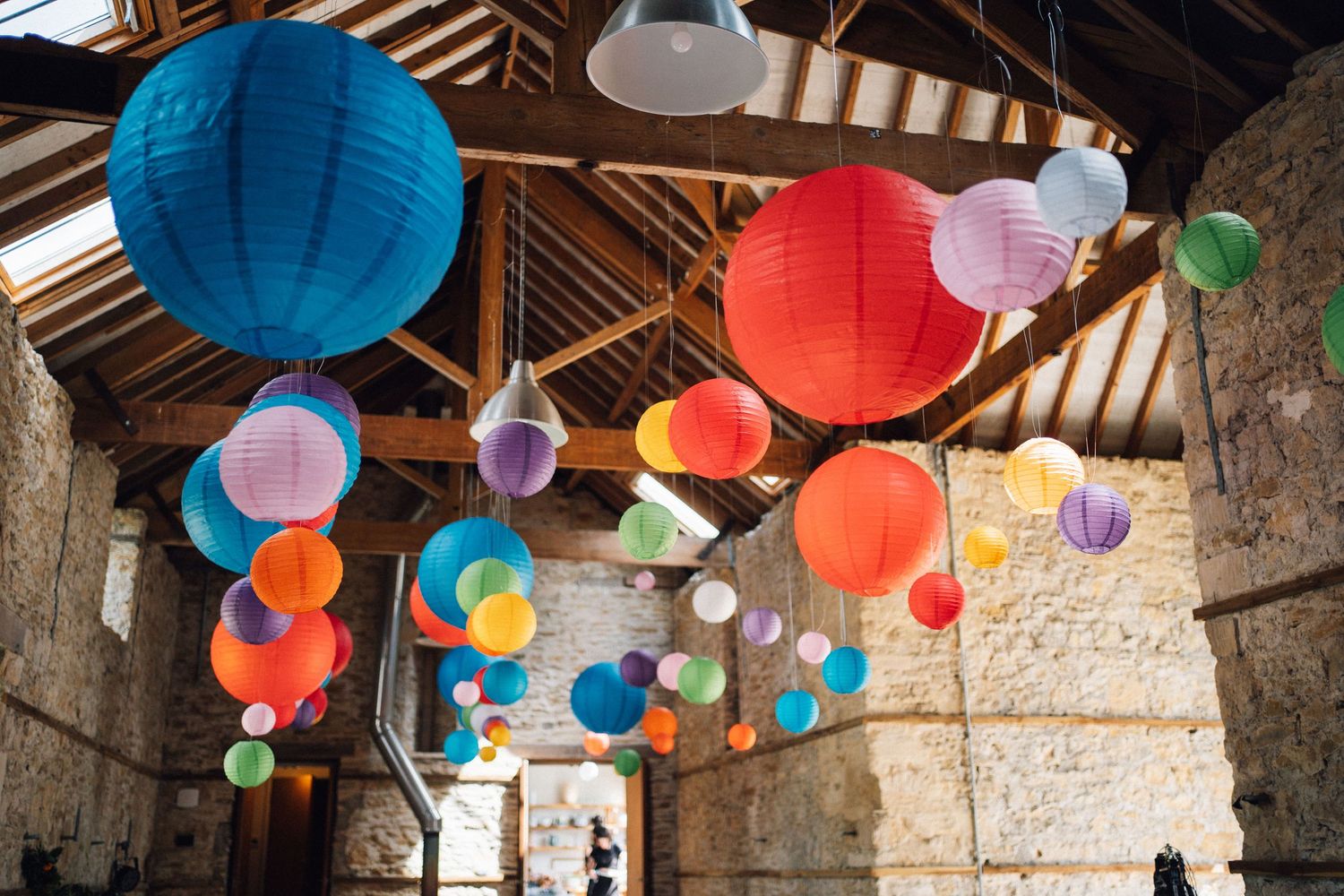 Colorful paper lanterns hanging from rustic barn ceiling in vibrant red, blue, green, and pink hues.