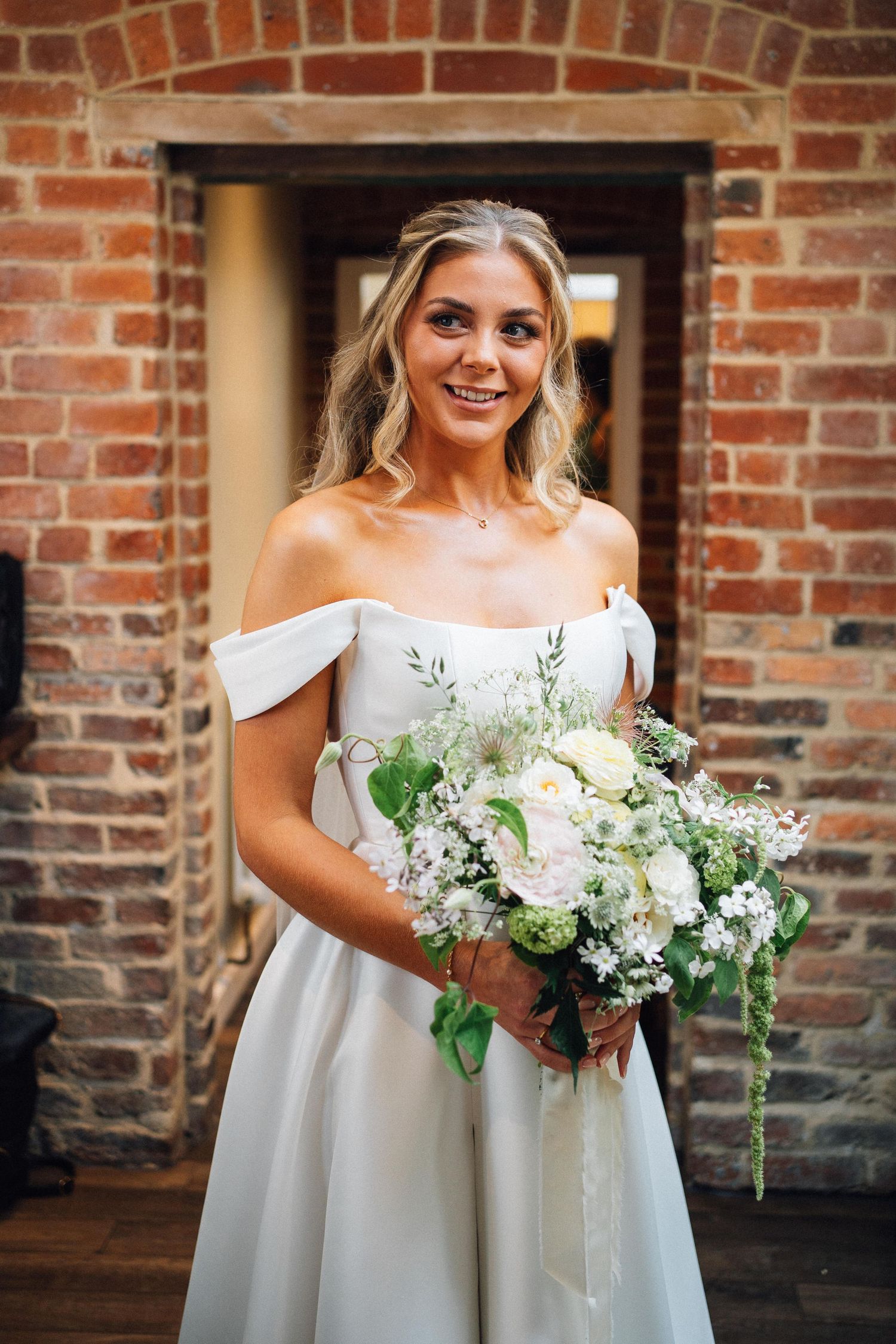A bride in an off-shoulder white gown holds a lush bouquet of white and green flowers against a brick wall backdrop.