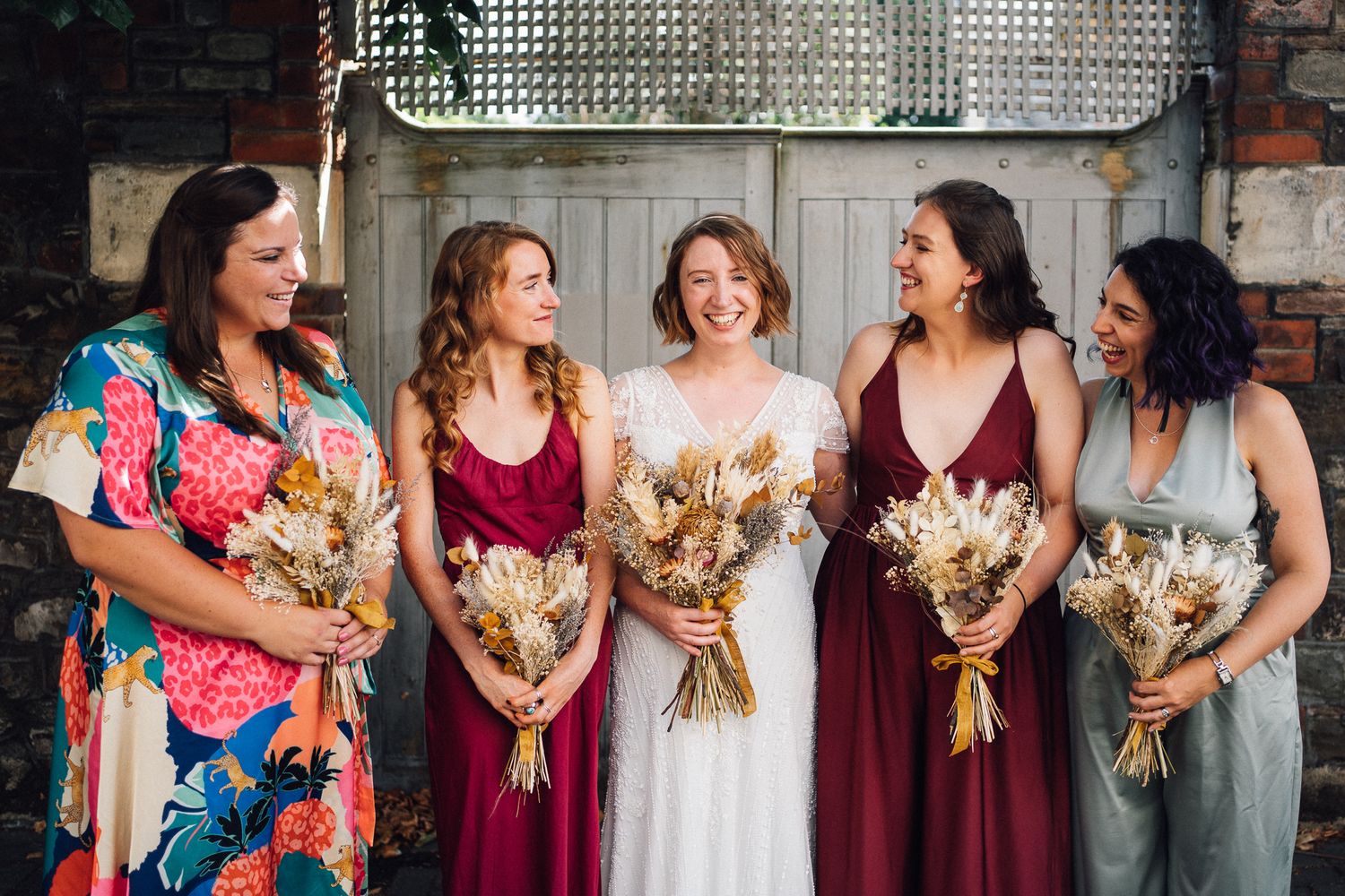 A row of bridesmaids in burgundy dresses and floral dresses stand together holding bouquets at a wedding.