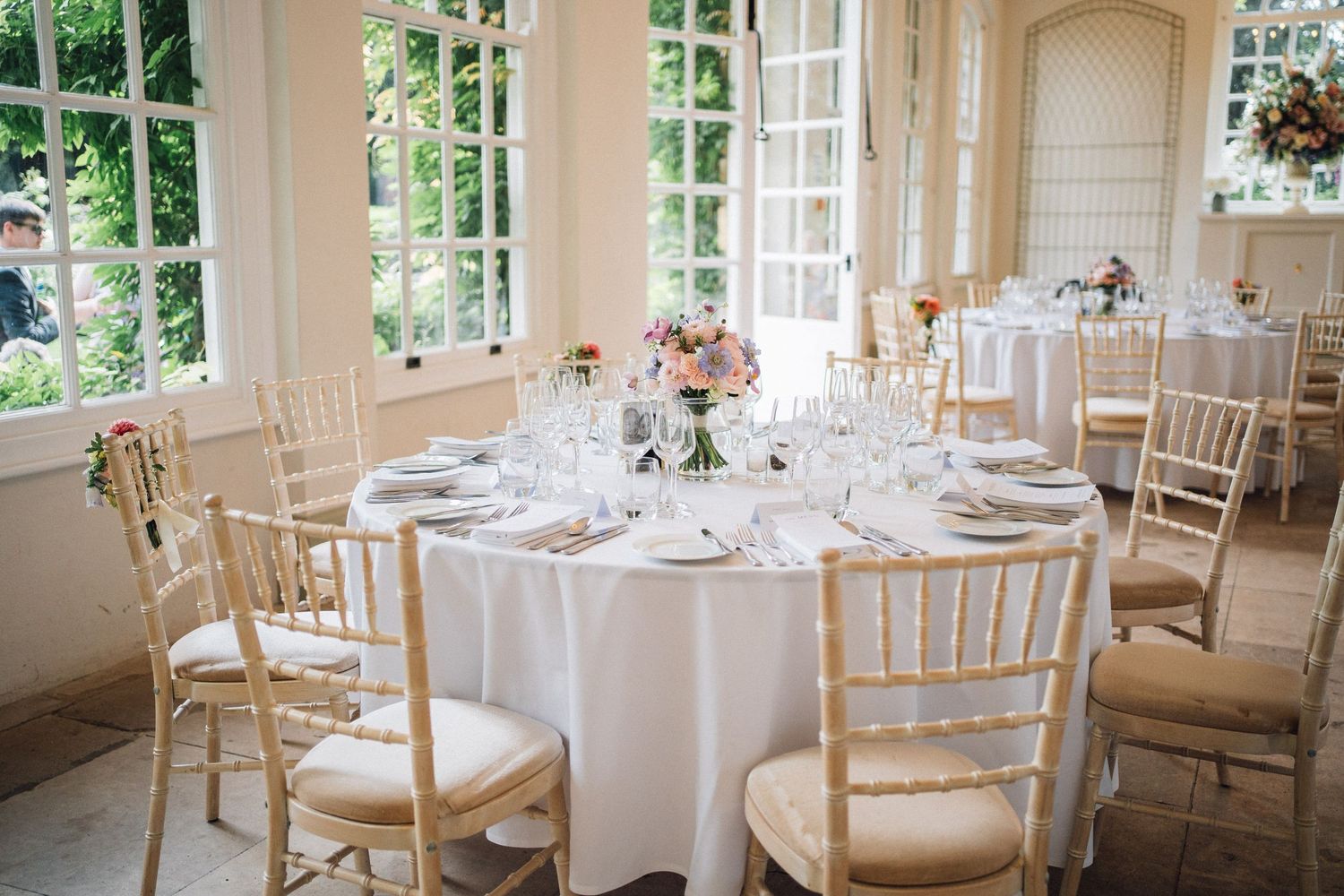 Elegant wedding reception table with white linens and chiavari chairs set in a bright sunlit conservatory room.