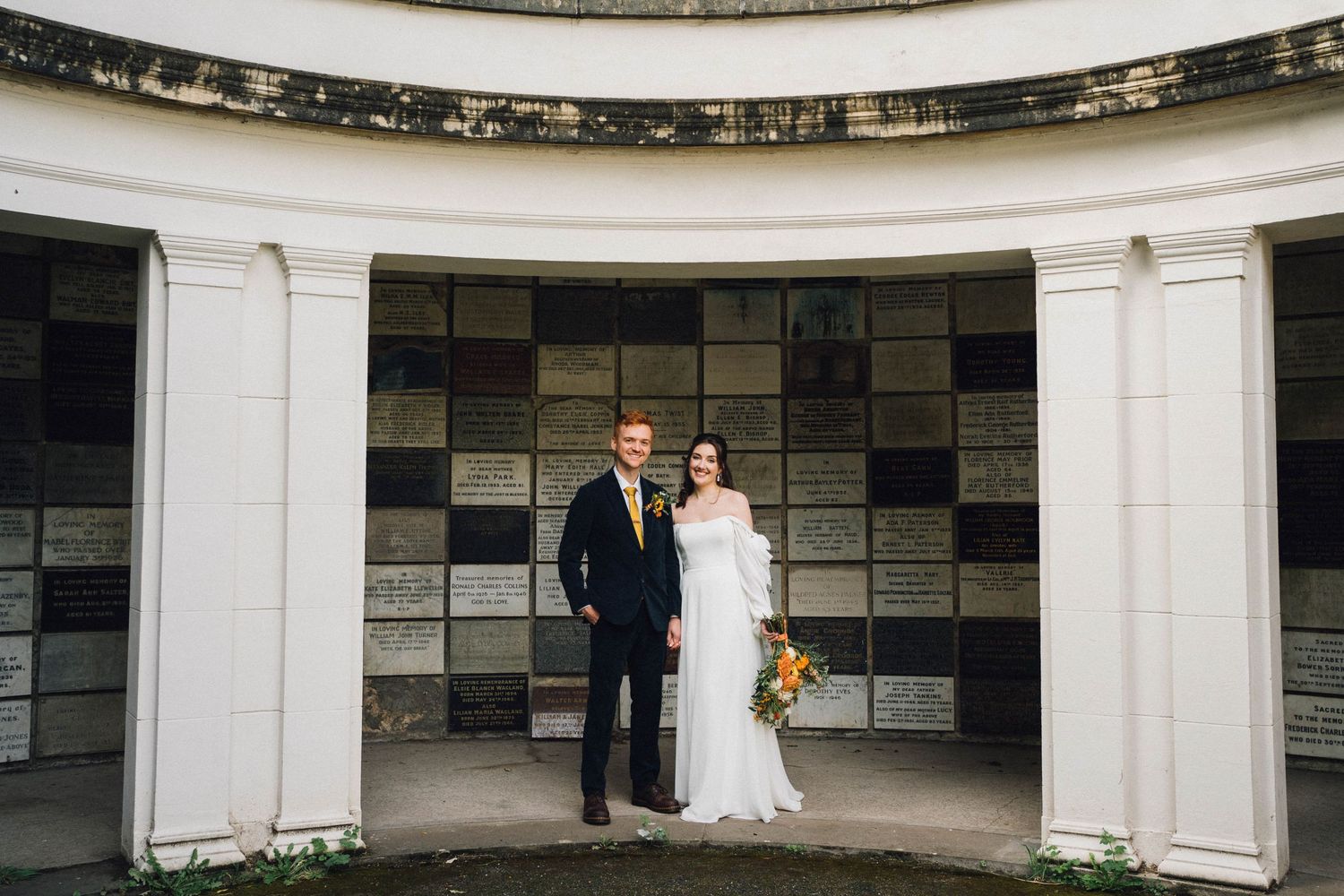 A wedding couple stands in a curved white colonnade walkway with stone walls creating an elegant architectural backdrop.