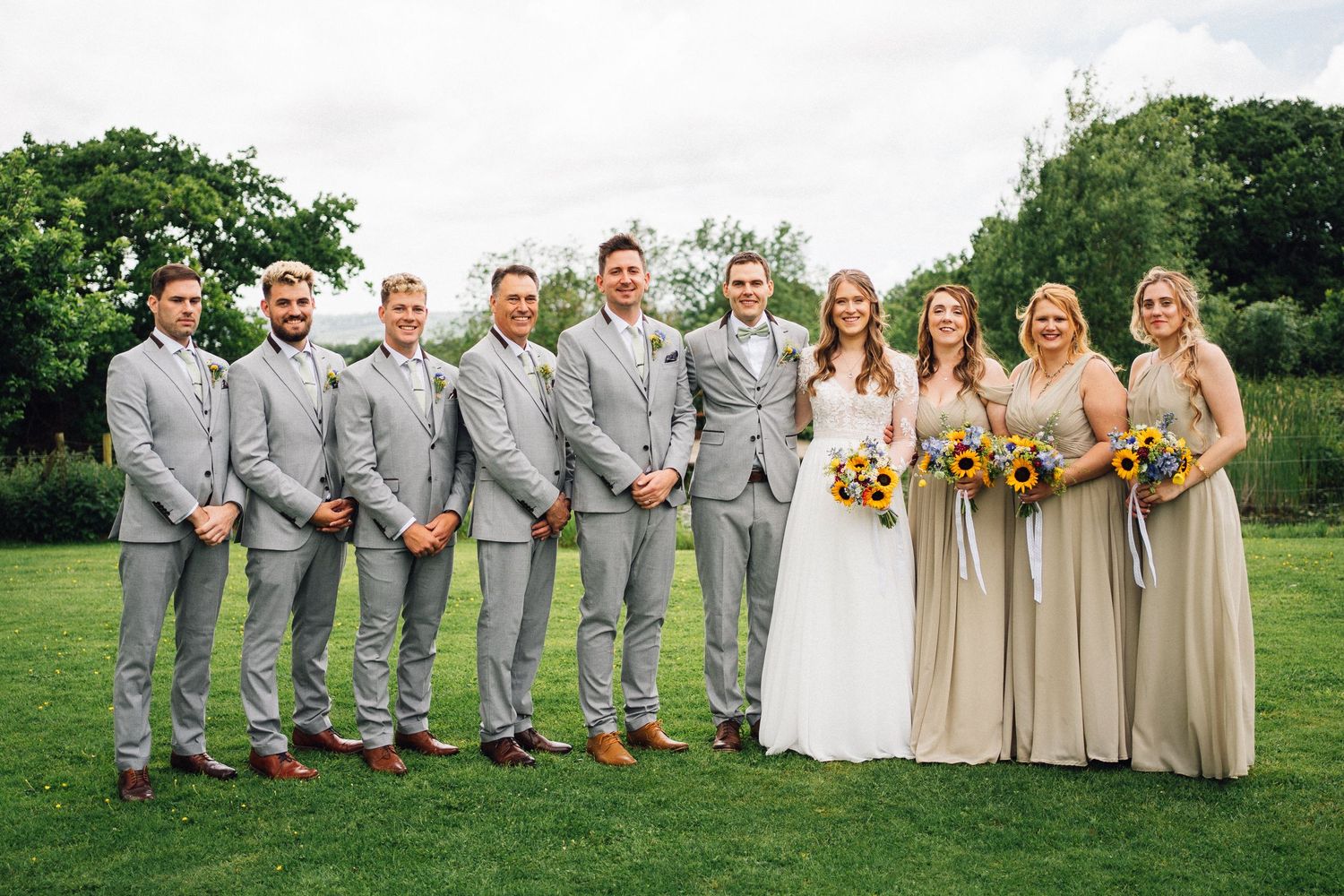Wedding party lined up outdoors wearing coordinated gray suits and neutral dresses for formal photos.