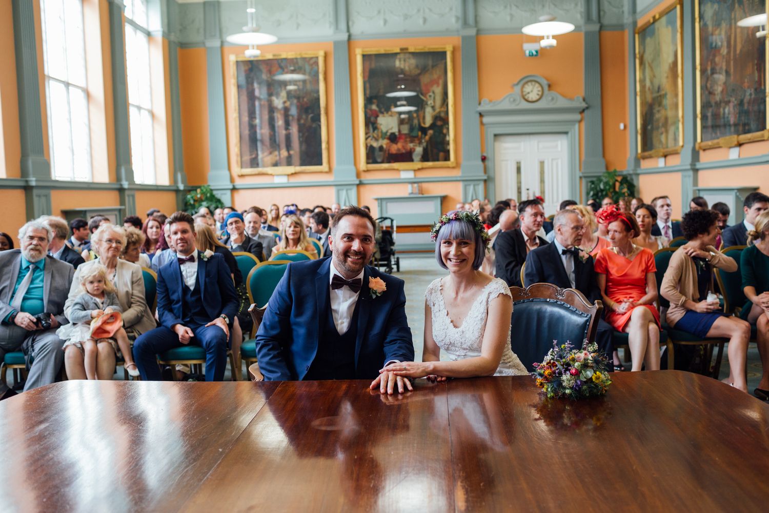 A wedding ceremony in a historic room with orange walls as the couple sits at a wooden table surrounded by guests.