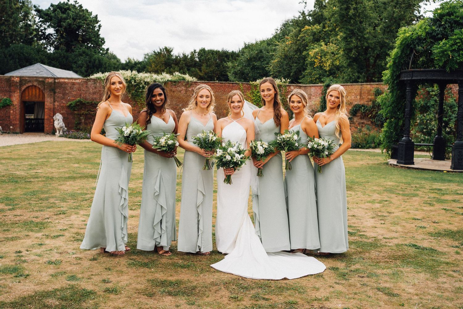 Wedding party stands outdoors in a garden setting with bridesmaids wearing sage green dresses and holding bouquets.