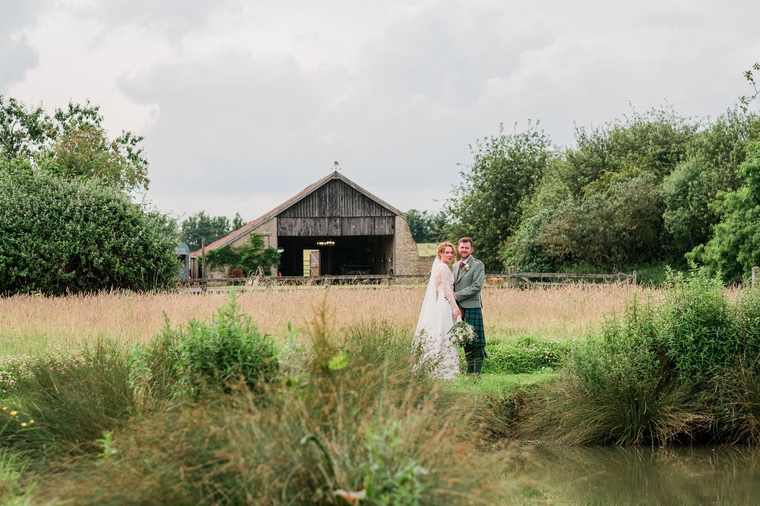 A rustic wooden barn provides a scenic backdrop for a couple in formal attire standing among tall grasses.