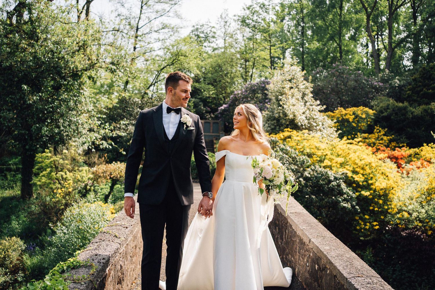 A couple walks along a scenic garden path surrounded by blooming yellow forsythia bushes and spring foliage.