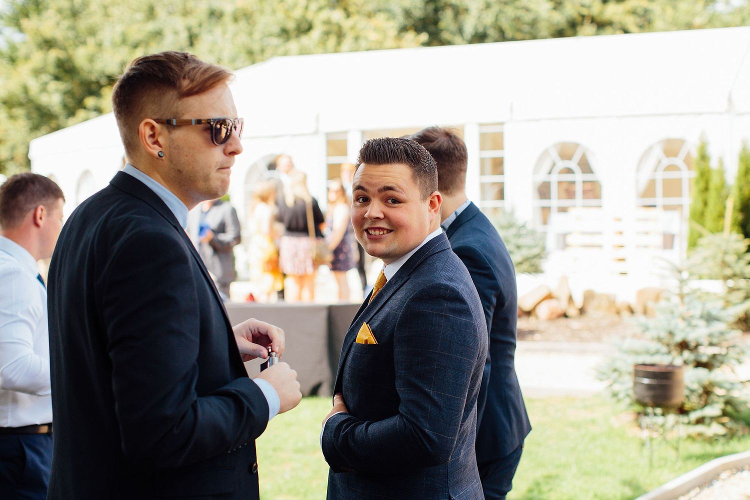 Wedding party members interact outdoors on a sunny day with greenery in the background.