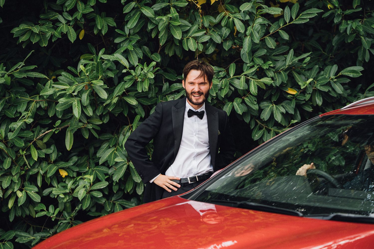 Men in formal attire standing near luxury cars and garden foliage at an outdoor event.