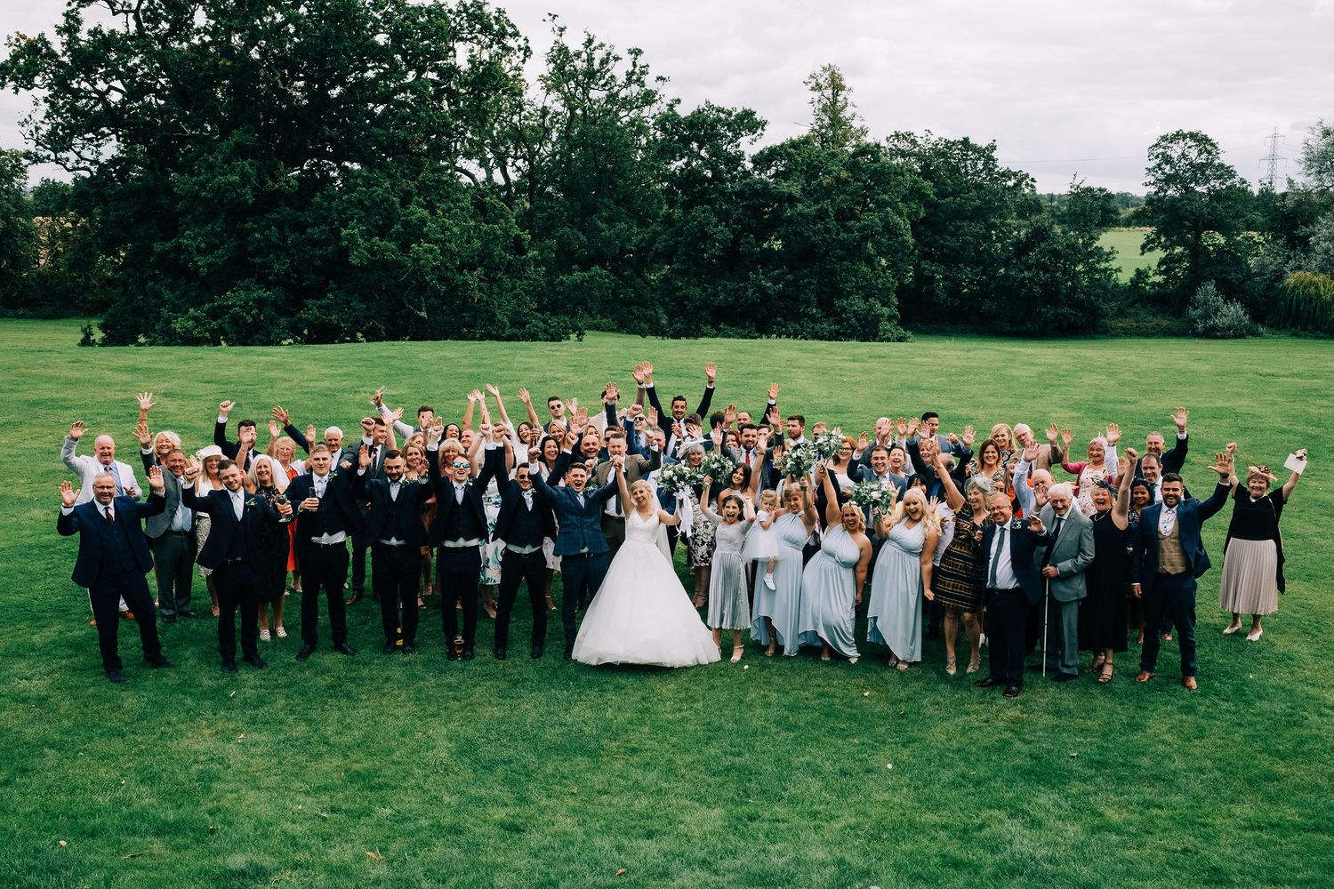Large group wedding photo with guests cheering on a green lawn.