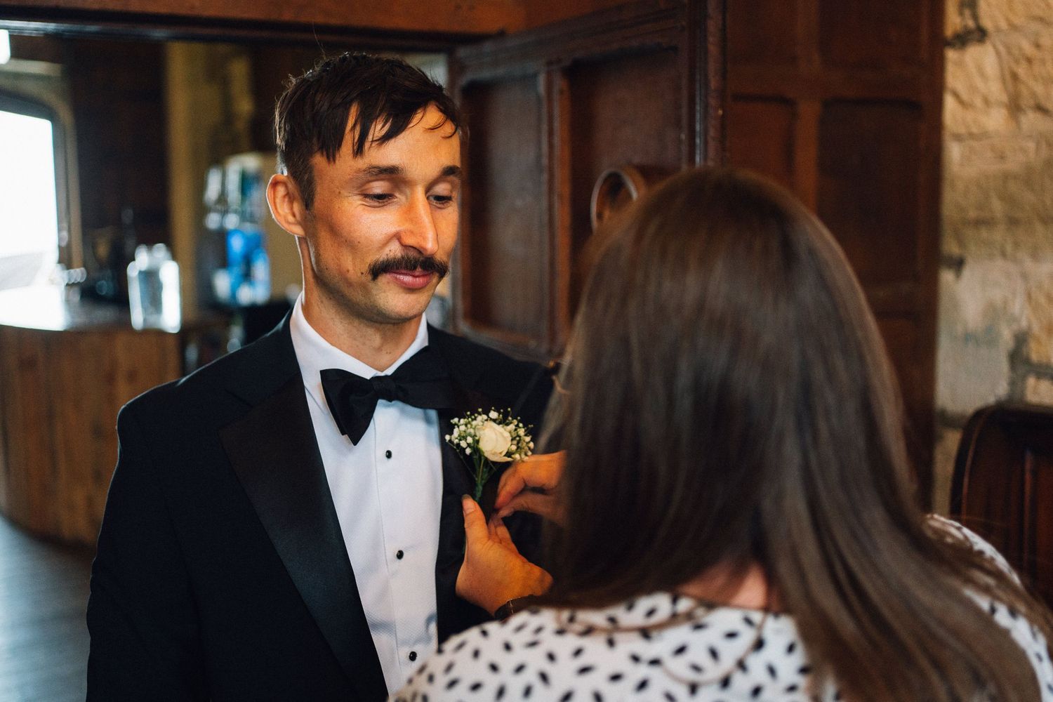 A person in a tuxedo and bowtie with a boutonniere speaks with someone indoors.