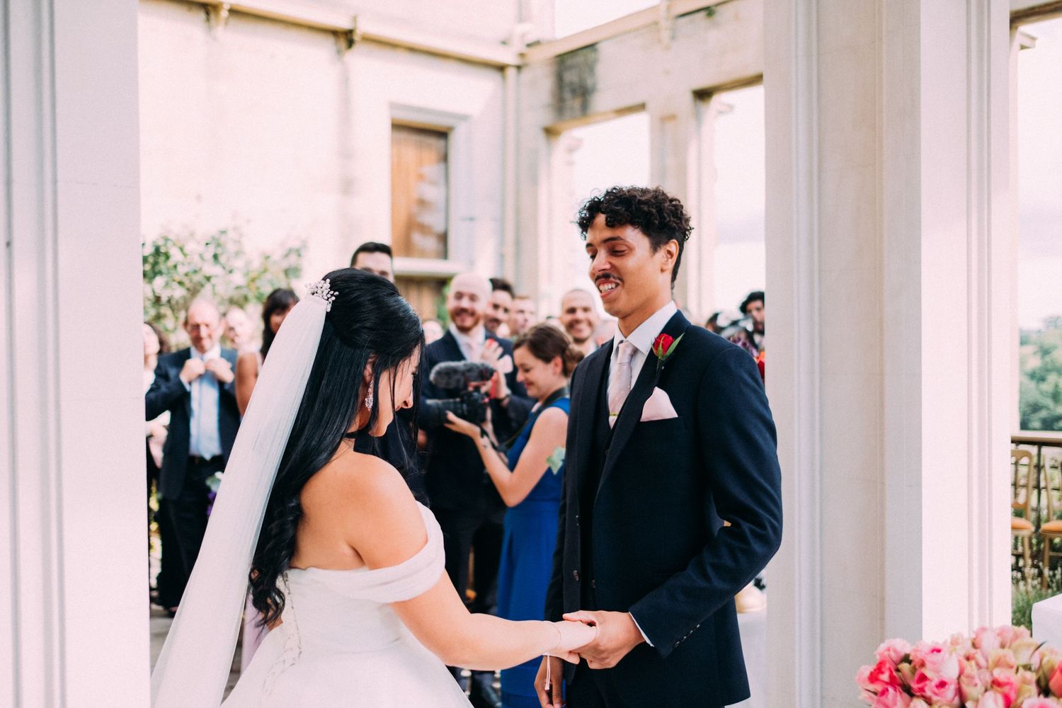 A couple exchanges rings during their wedding ceremony surrounded by white columns and pink flowers.