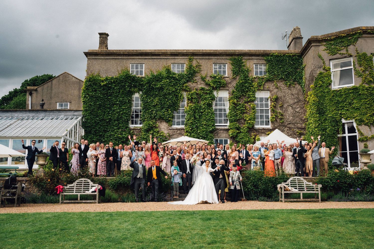 Large wedding gathering poses in front of an elegant ivy-covered stone building.