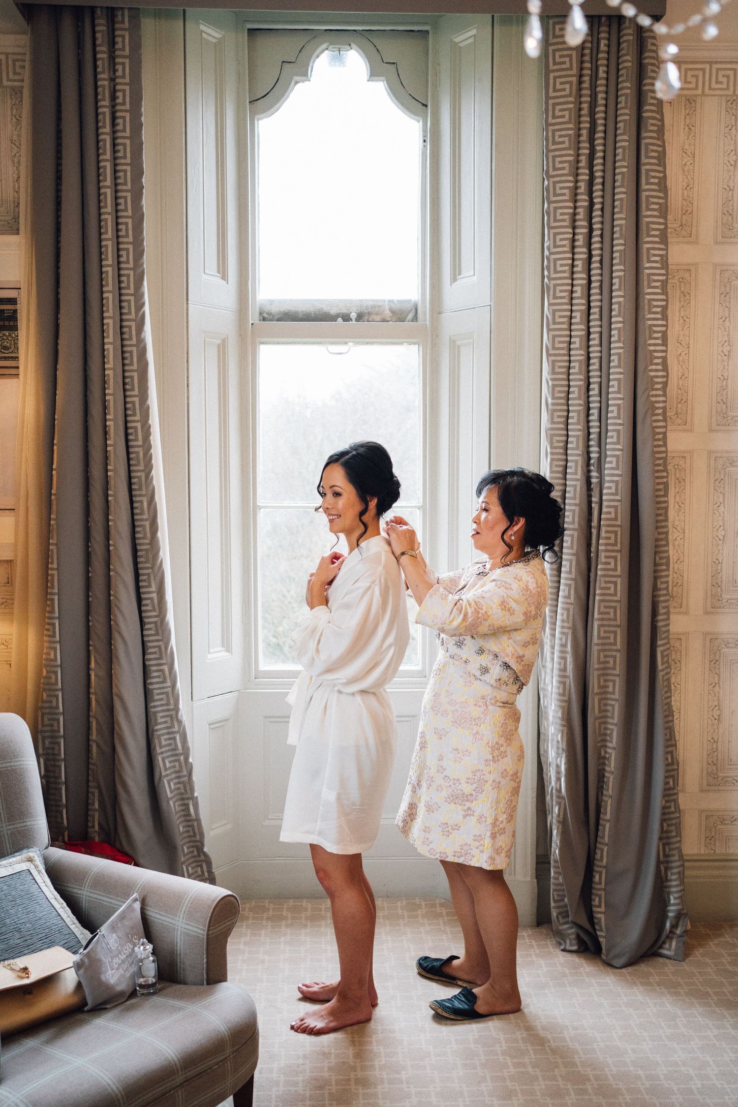 Playful candid moments during wedding preparation in a room with tall windows and gray curtains.