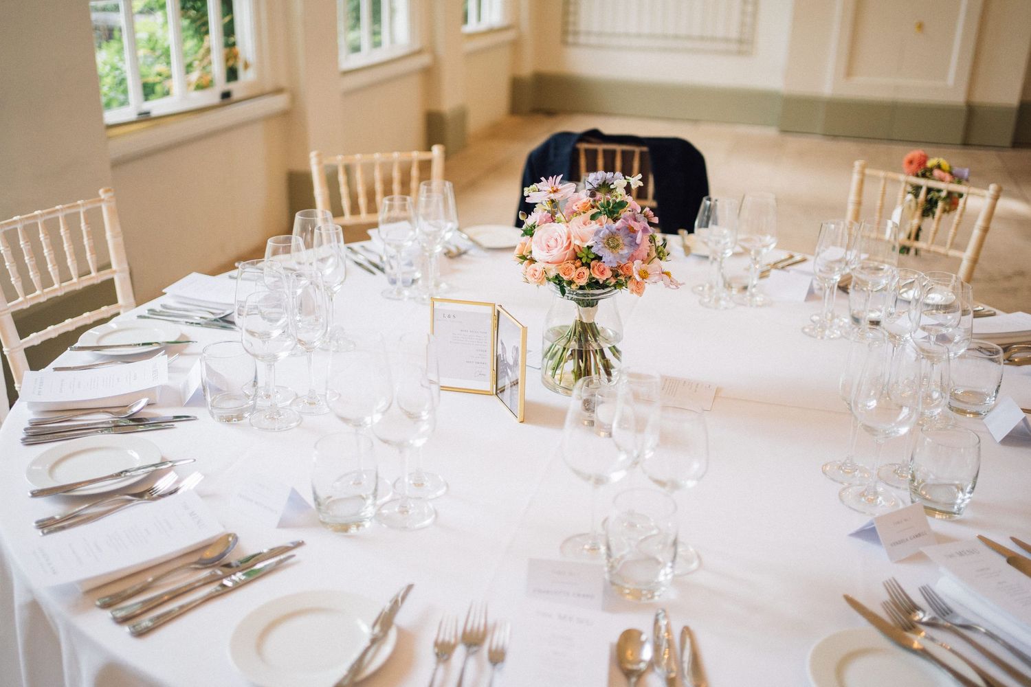 Elegant wedding reception tables with white linens, glassware and place settings in a bright room.
