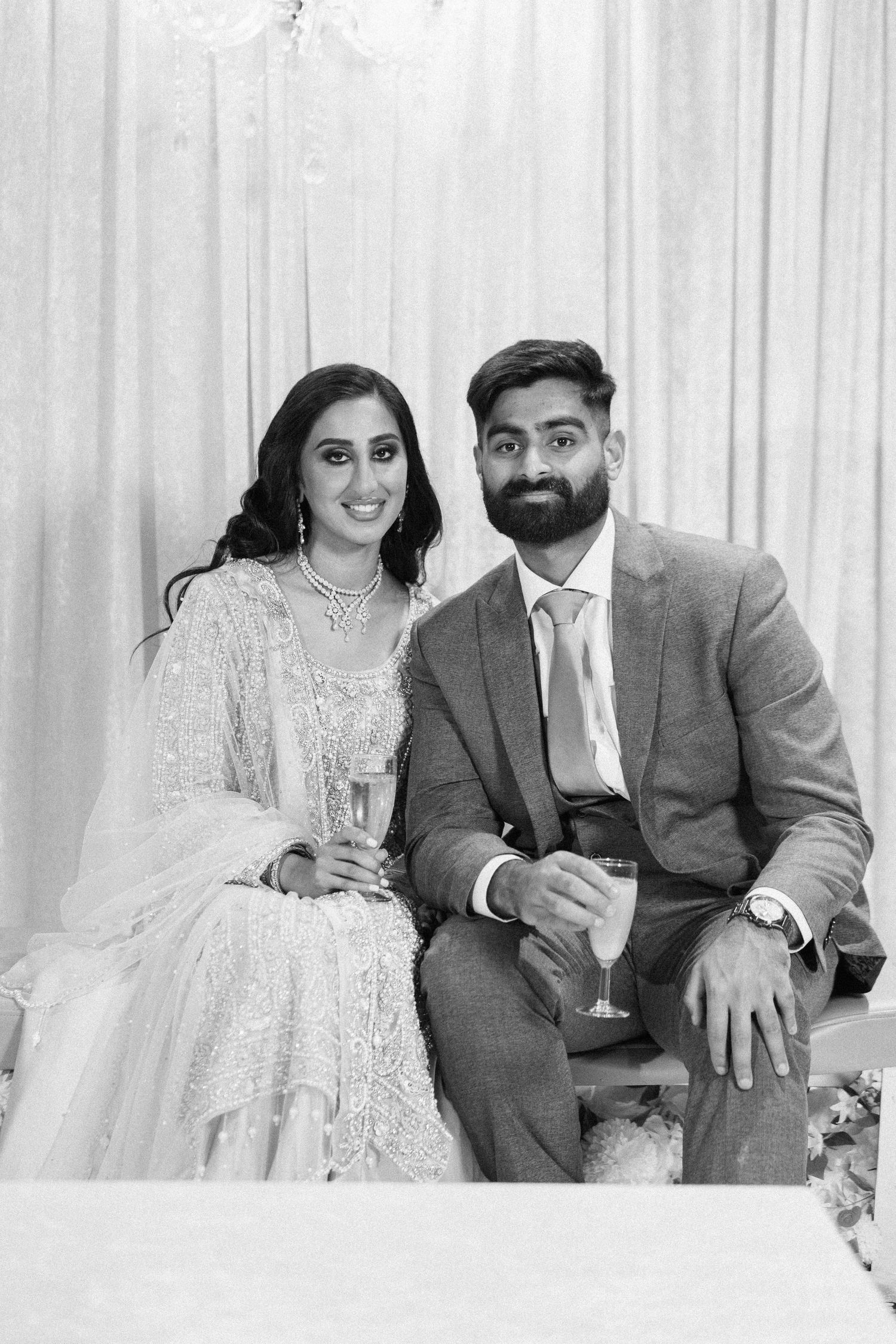 A couple in wedding attire share a celebratory drink together in a black and white portrait.