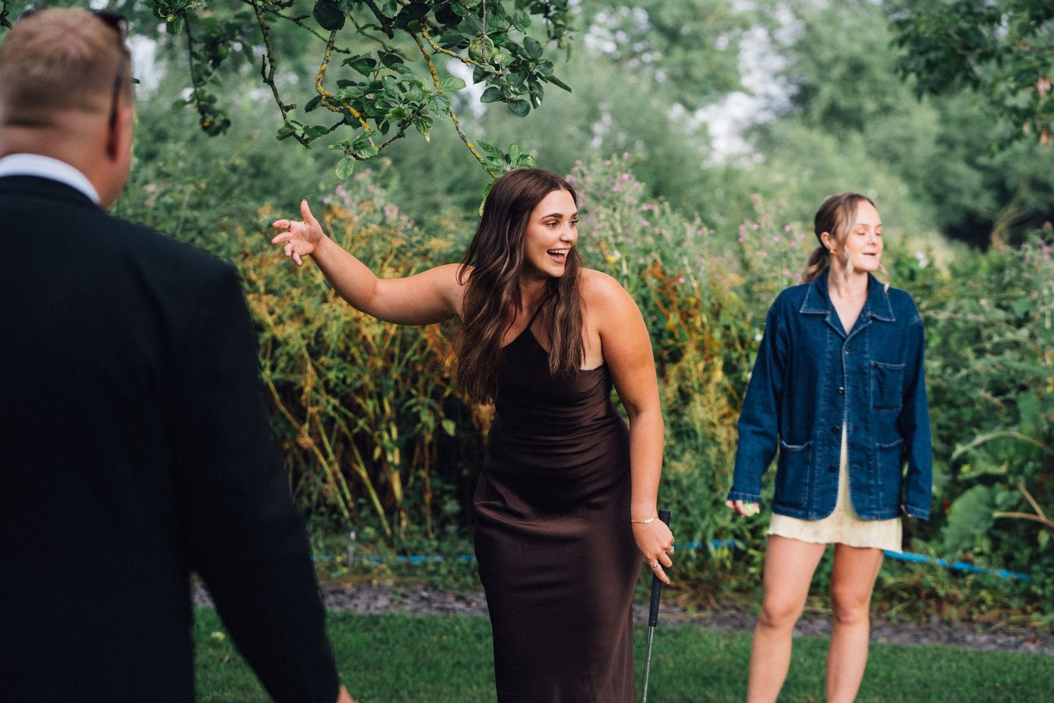 People interact outdoors in a garden setting with one wearing a brown dress and another in a blue denim jacket.