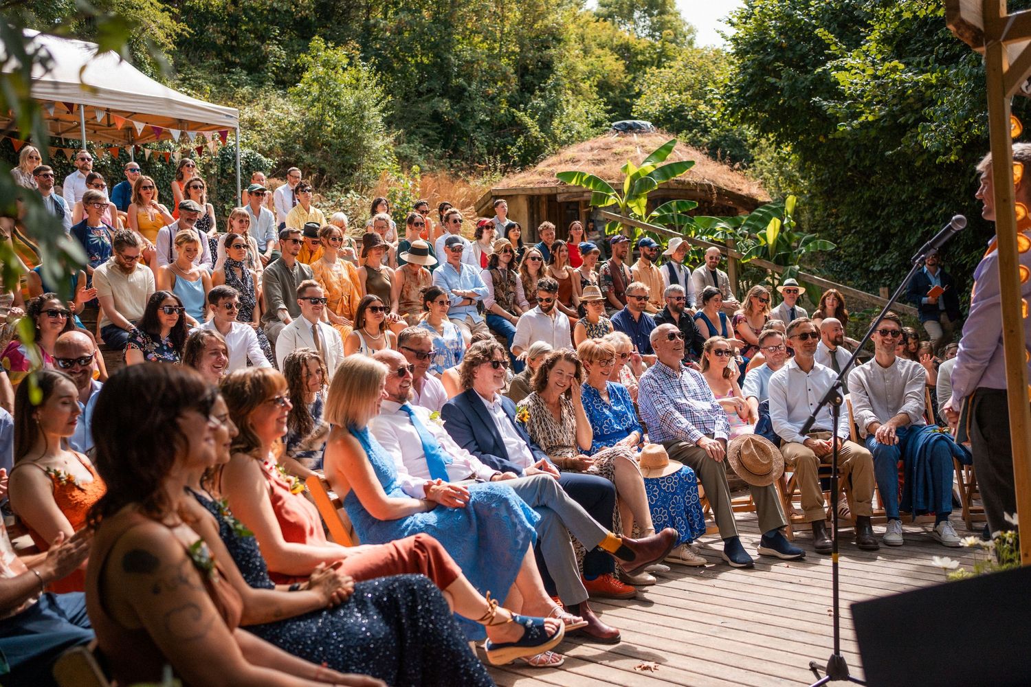 Large crowd gathered outdoors at a sunny garden event, seated on wooden steps, surrounded by lush greenery.