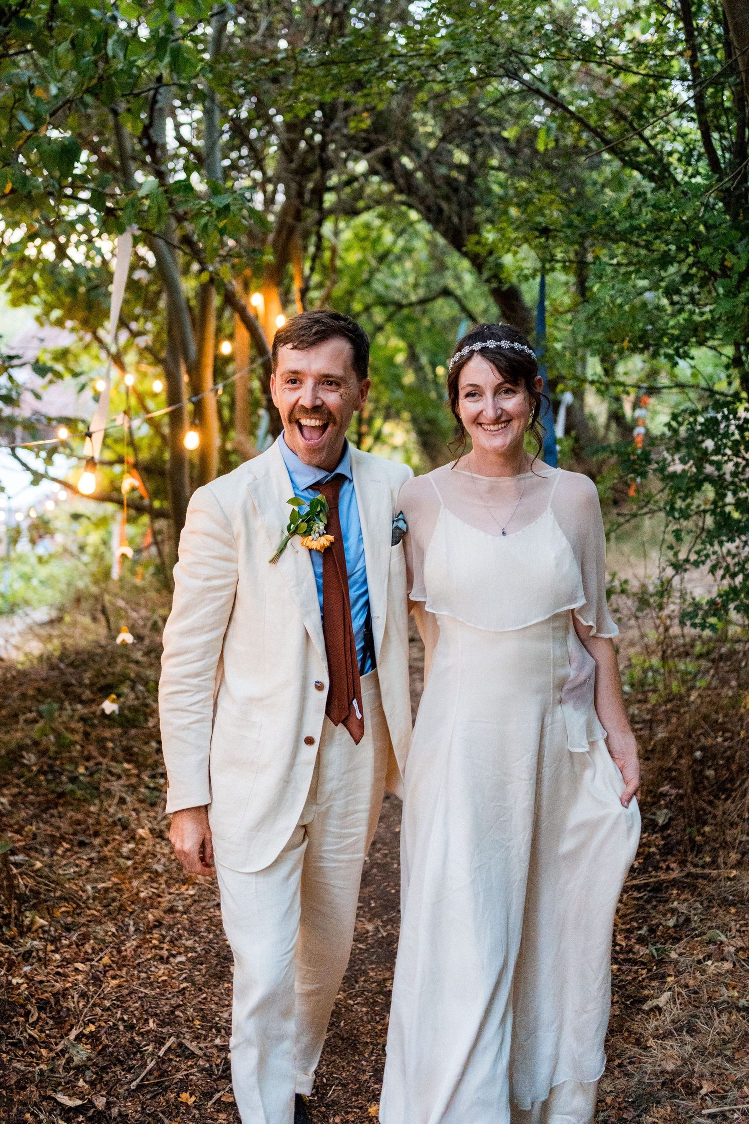 Happy couple in wedding attire posing outdoors under tree canopy with string lights, groom in cream suit, bride in white dress.