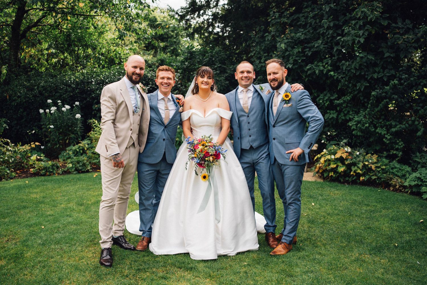 Wedding party members pose together on a lush green lawn during an outdoor summer celebration.