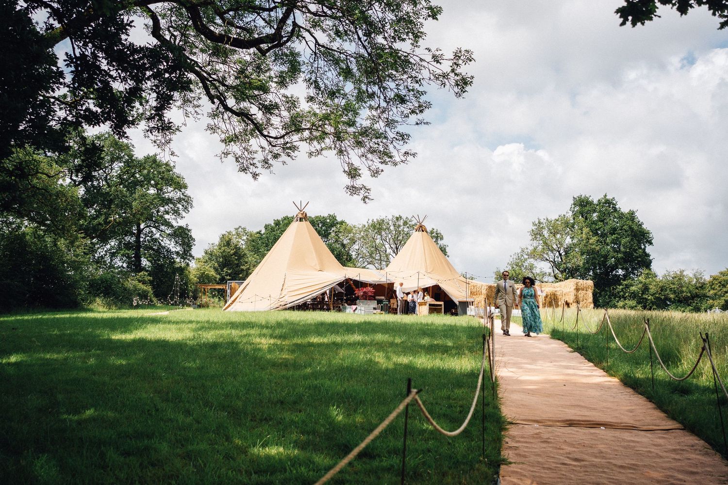 A panoramic view of glamping tents and teepees set up in a lush green field with trees in the background.
