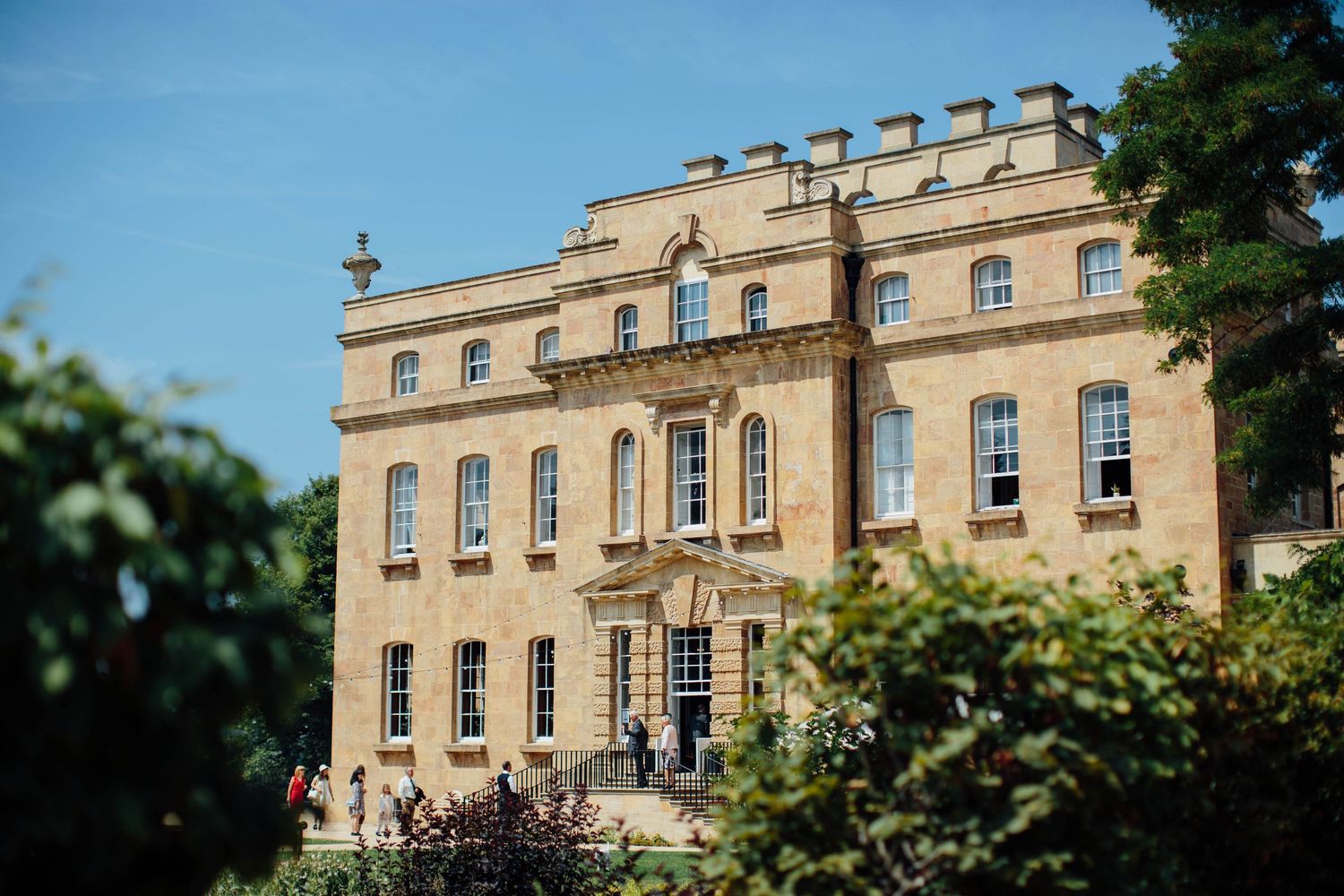 Grand Georgian stone manor house surrounded by lush green trees under a bright blue summer sky.