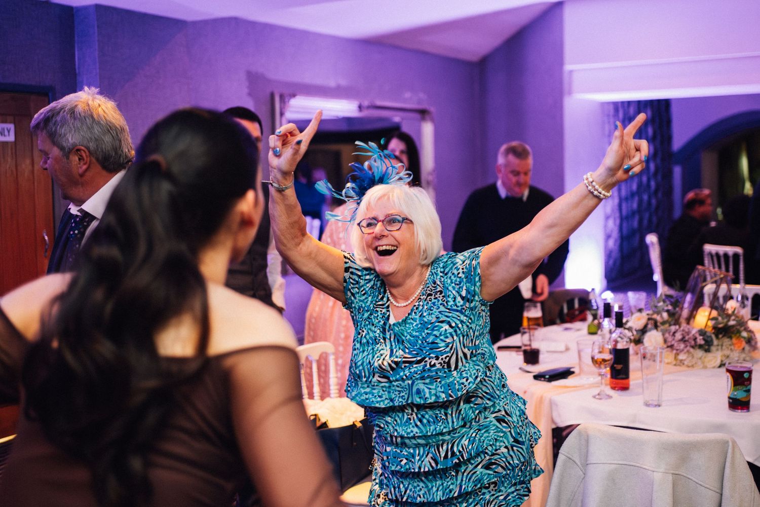 Wedding reception guests dancing and celebrating under purple mood lighting in an indoor venue.