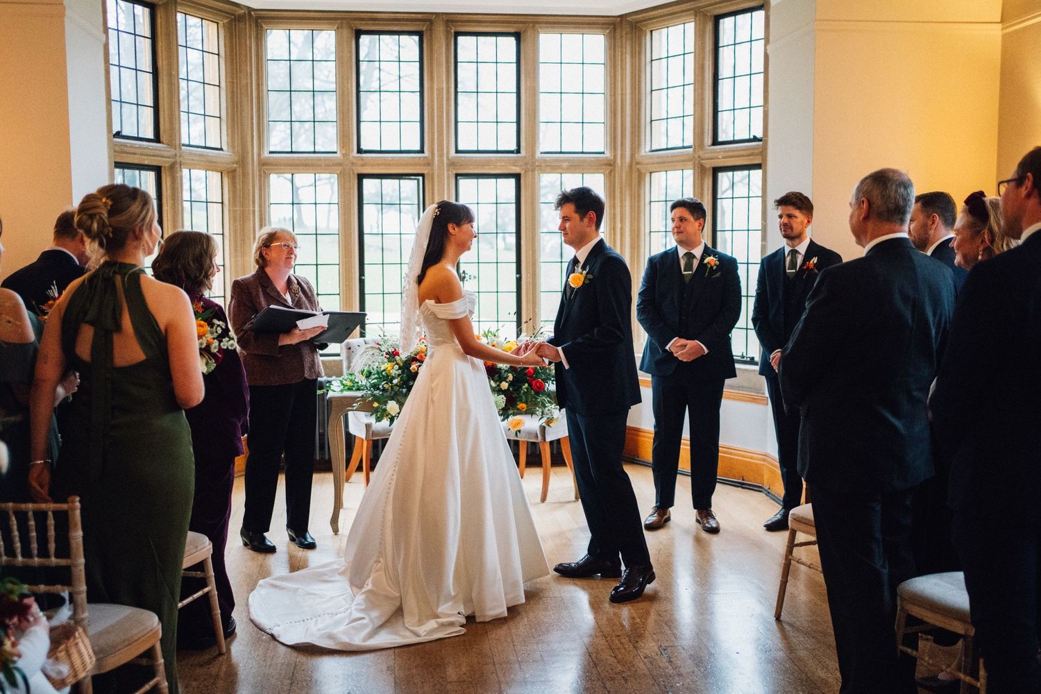 Wedding party stands in elegant room with large windows during indoor celebration.