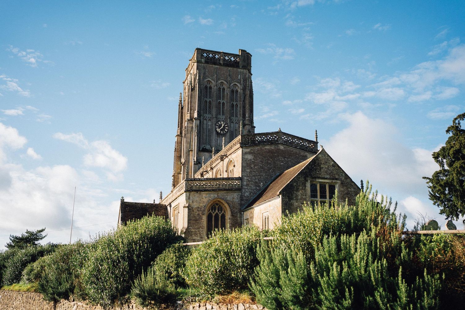 Historic stone church with tall Gothic tower surrounded by lush green shrubs under a blue sky with white clouds.