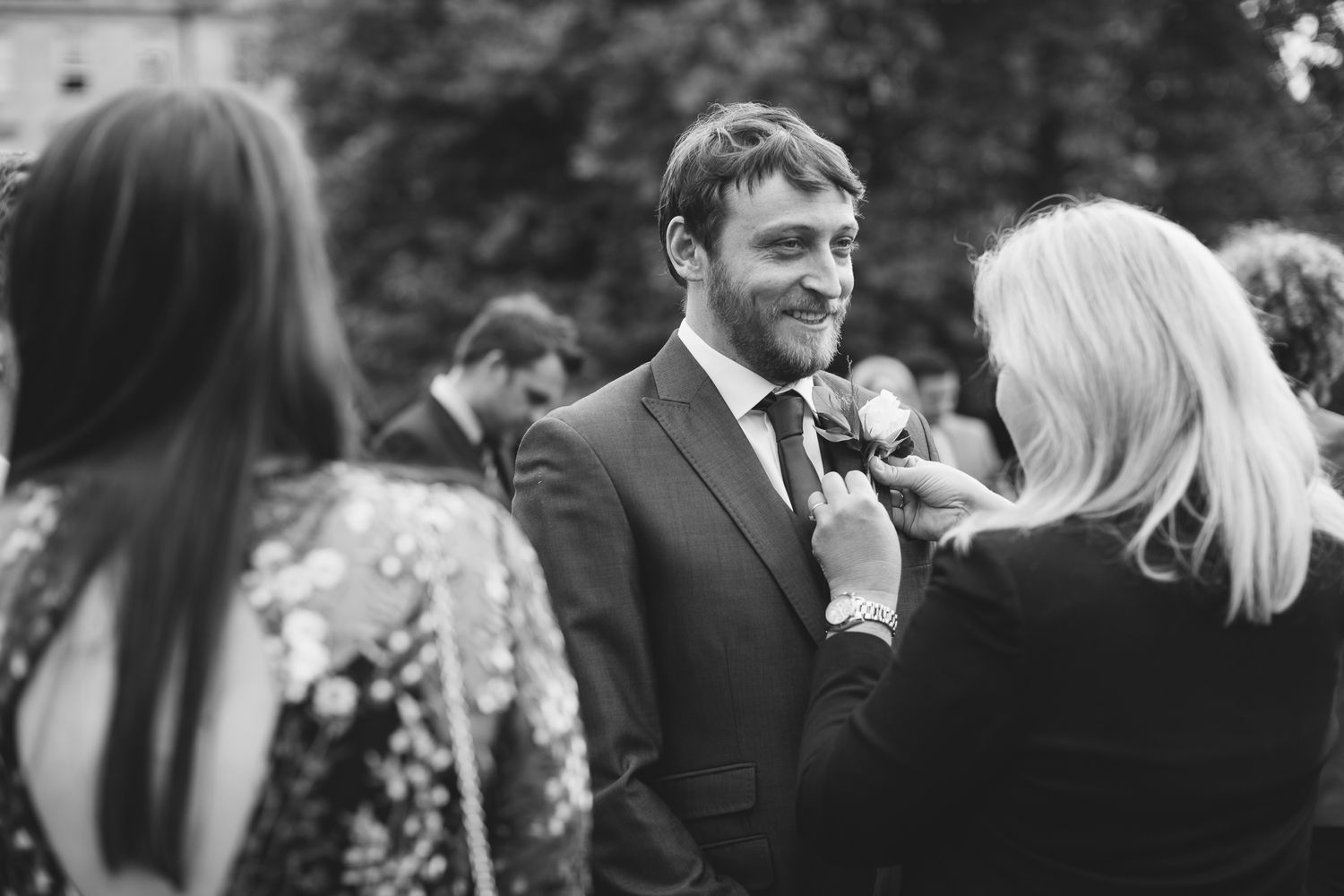 Black and white wedding photo of groom smiling as woman pins boutonniere on his suit lapel.