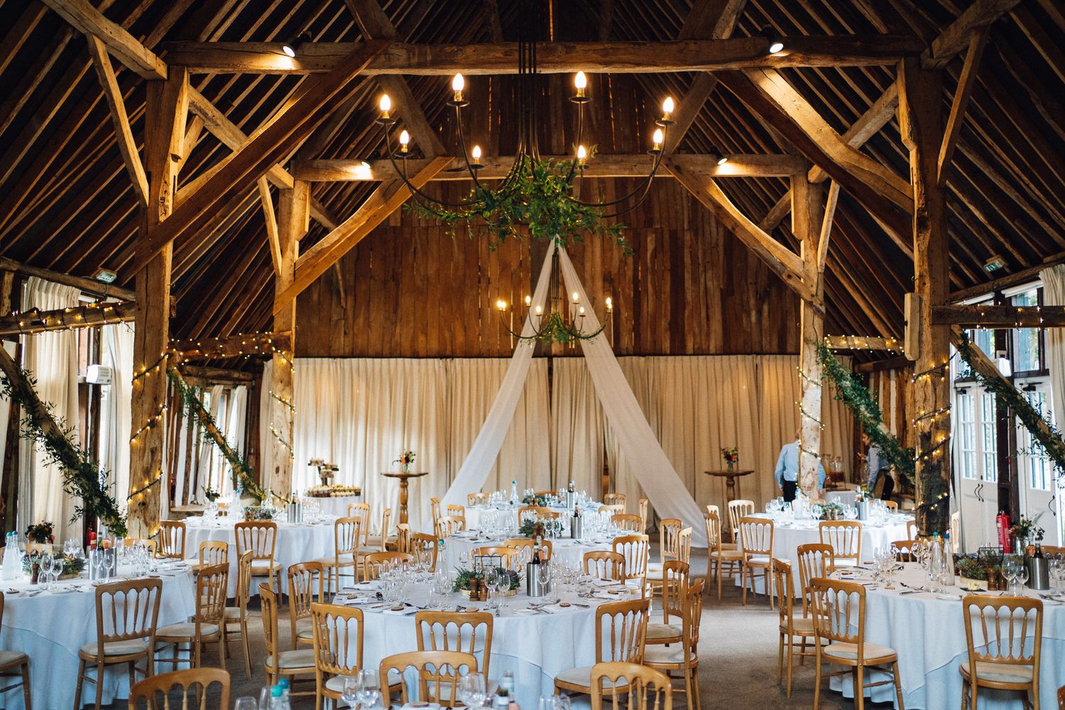 Rustic barn wedding reception venue with wooden beams and elegant white table settings illuminated by hanging lights.