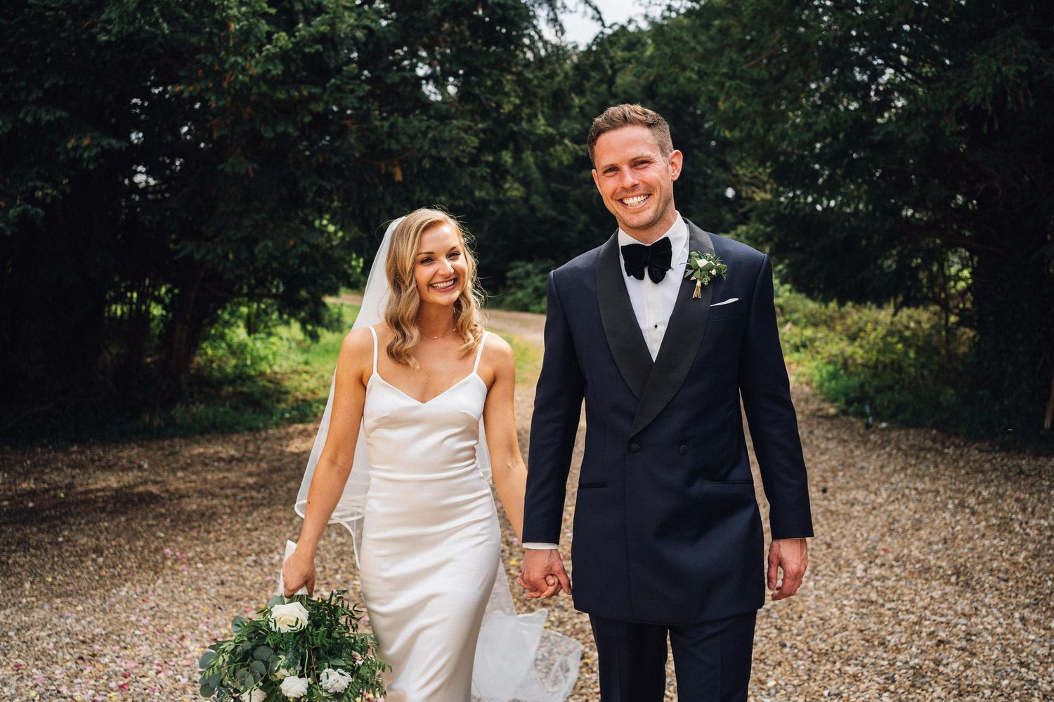 Newlywed couple in formal attire poses together on a rustic outdoor path surrounded by greenery.