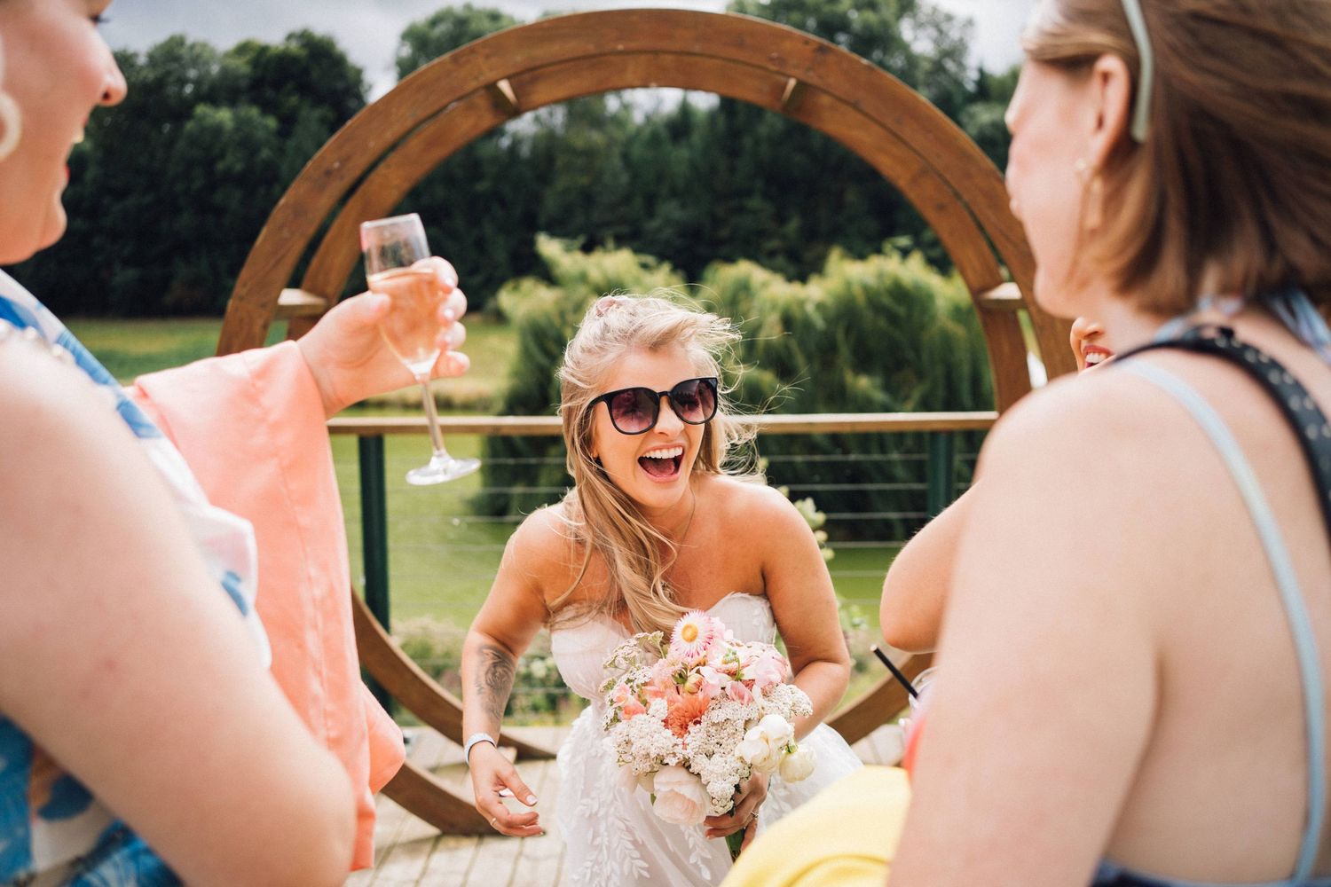 A group of wedding guests laugh together while seated at an outdoor reception table in a garden setting.