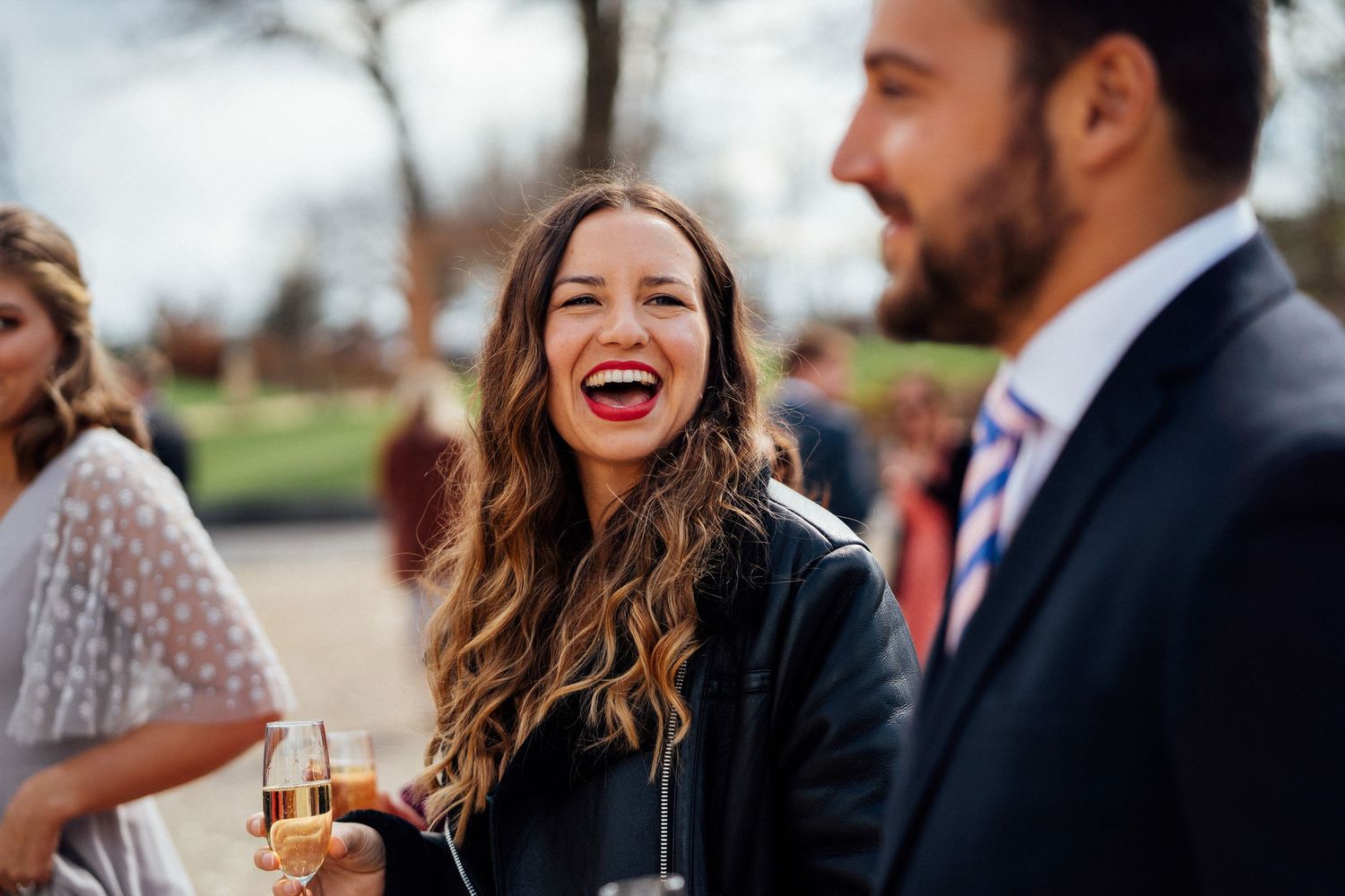 A woman with long wavy hair laughs joyfully while holding a drink at an outdoor social gathering.
