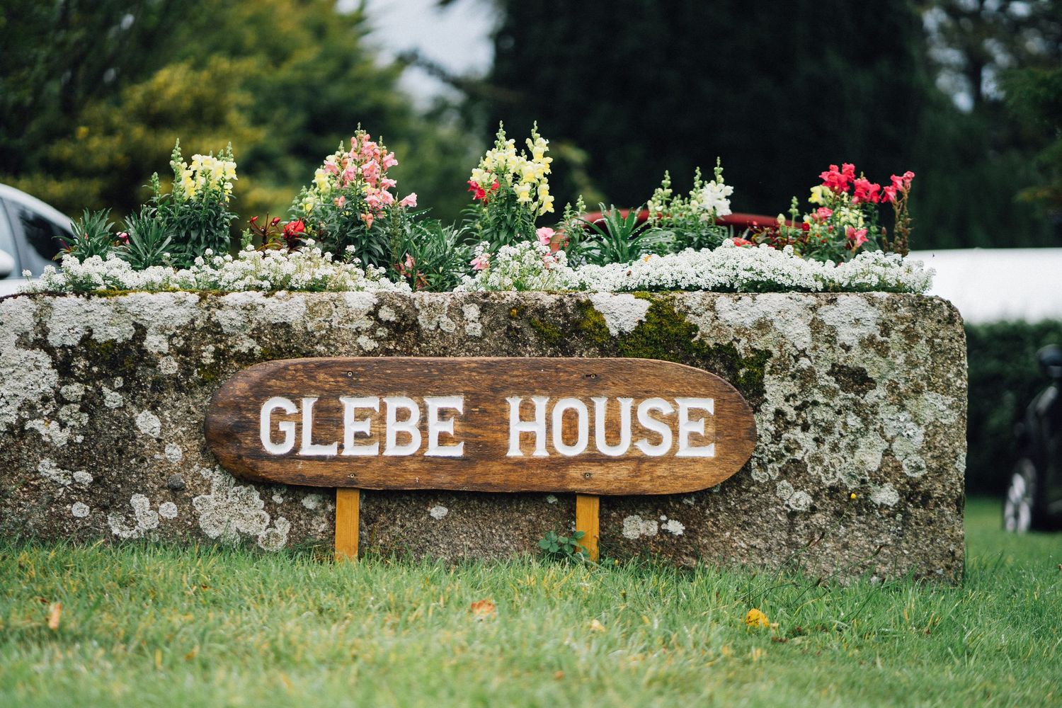 Stone wall with rustic 'Glebe House' signage surrounded by greenery and flowers in a garden setting.